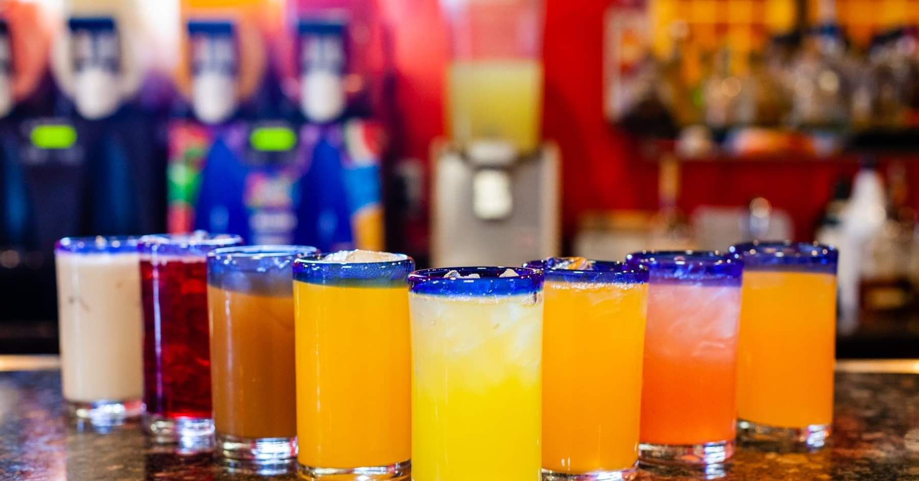 Colorful cocktails in glasses with blue rims, arranged on a bar. Frozen drink dispensers are visible in the background.