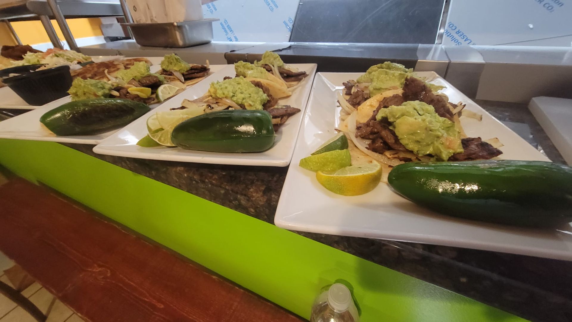 Tacos topped with guacamole, served with lime and jalapeño on white plates, prepared on a countertop.