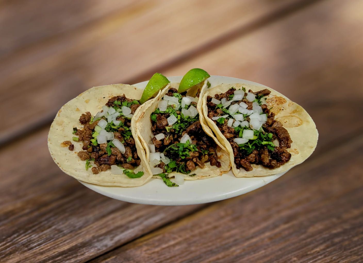 Three tacos on a white plate with meat, onions, cilantro, and lime wedges, set on a wooden surface.