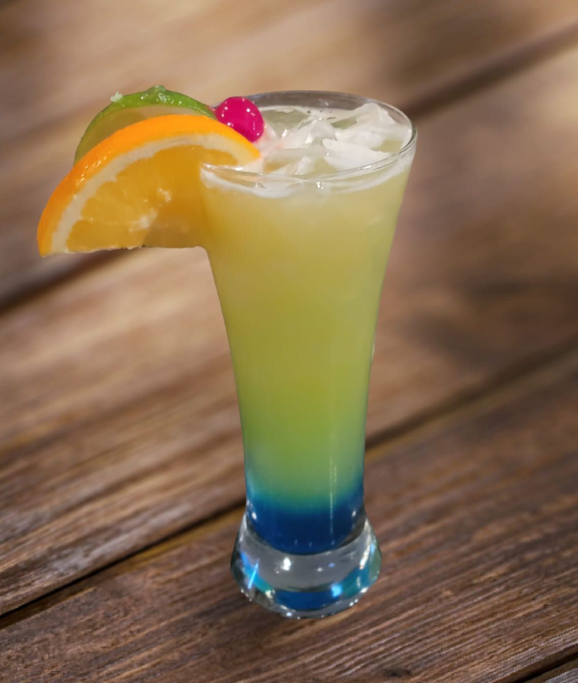 Cocktail in a tall glass, layered blue, green, and yellow, garnished with orange slice, cherry, and lime, on wood.
