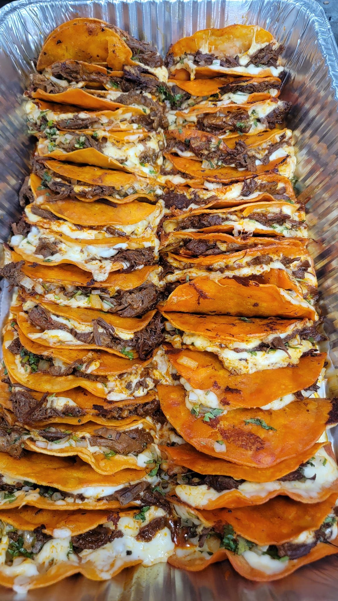 Birria tacos in a foil tray, arranged in rows. Orange-colored tortillas filled with meat and cheese.