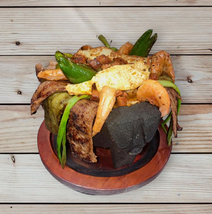 Molcajete dish: Grilled meats, vegetables, and shrimp in a stone bowl on a wooden stand.