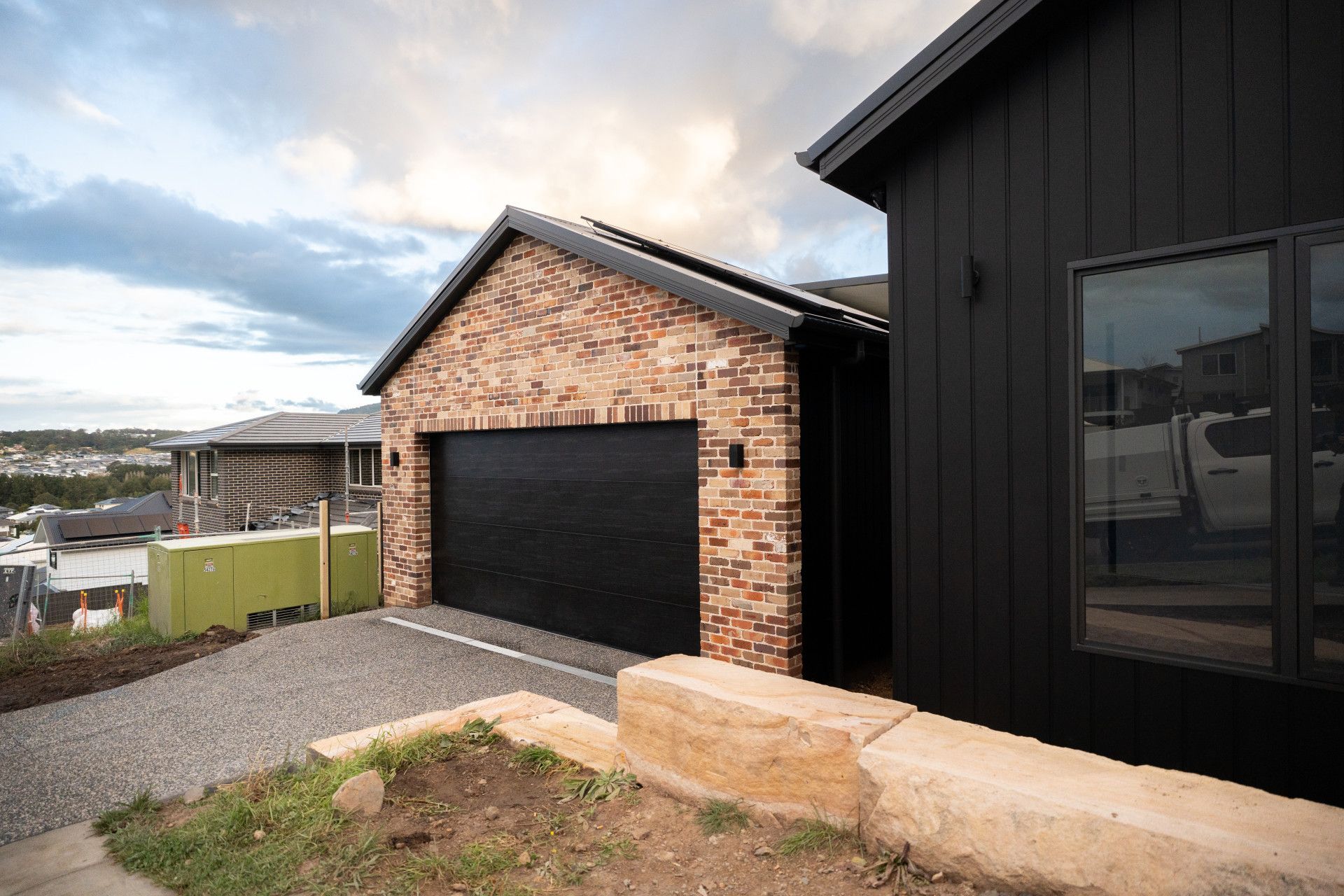 Modern house exterior with brick garage, black siding, and gravel driveway under a cloudy sky - Matt Made Building - Builder Camden, NSW.
