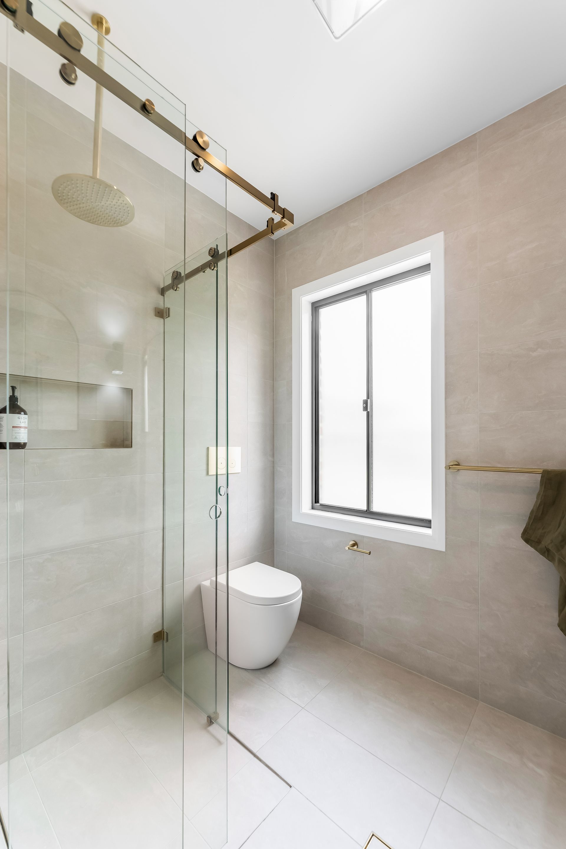Modern Bathroom With Glass Shower, White Toilet, and Window on Beige Tile Walls — Matt Made Building in Camden, NSW