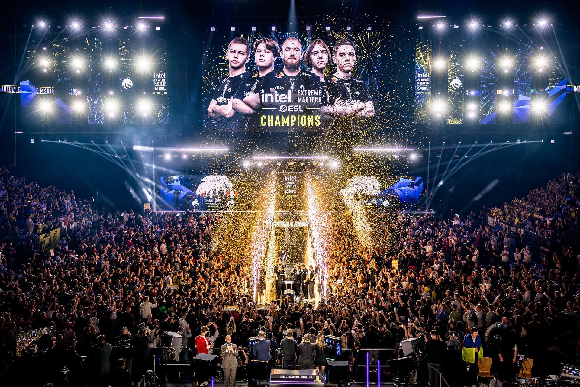 A crowd cheers as a victorious esports team is celebrated onstage, confetti falling, bright lights.