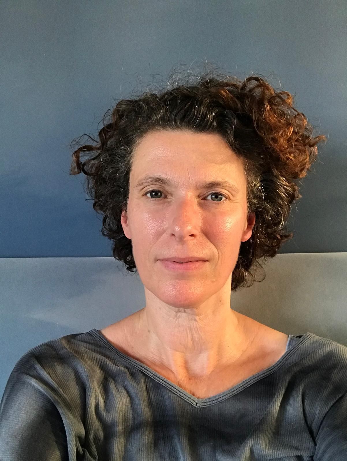 Vrouw met krullend bruin haar, gekleed in een donkergrijze blouse, tegen een blauwgrijze achtergrond, met een lichte glimlach.
