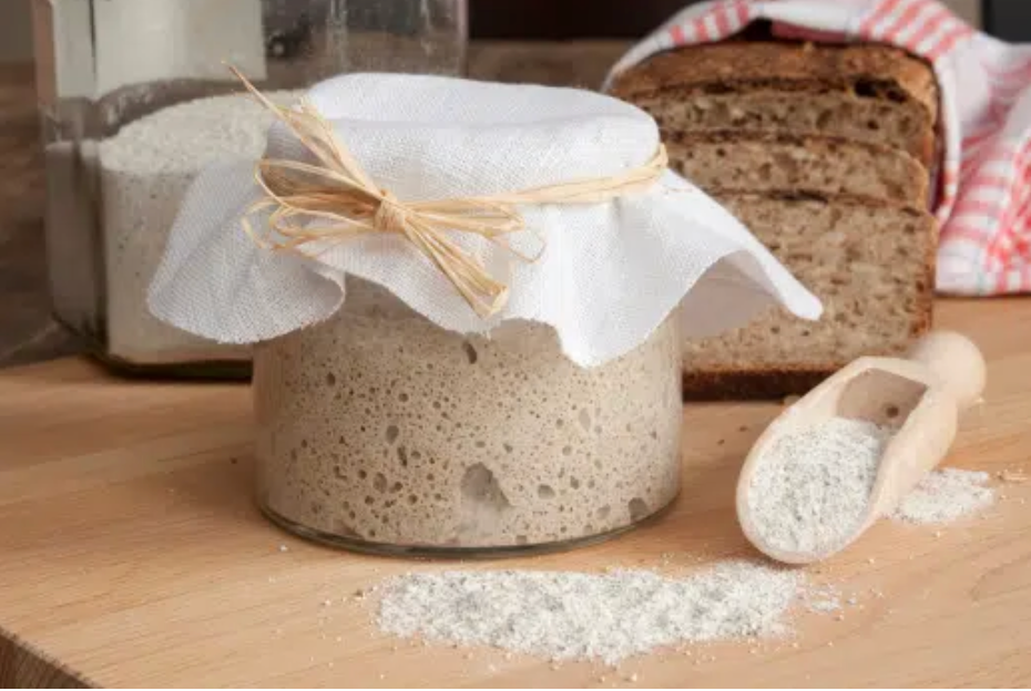 Een zuurdesemstarter in een pot, afgedekt met een doek en vastgebonden met touw. Ook aanwezig zijn sneetjes brood, bloem en een houten ijsschep.