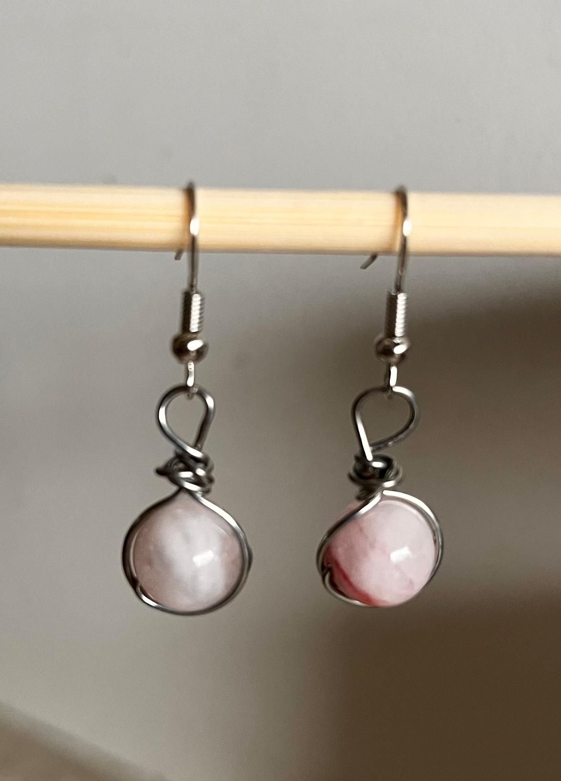 Een paar zilveren oorbellen met ronde, lichtroze hangers die aan een houten staafje hangen.