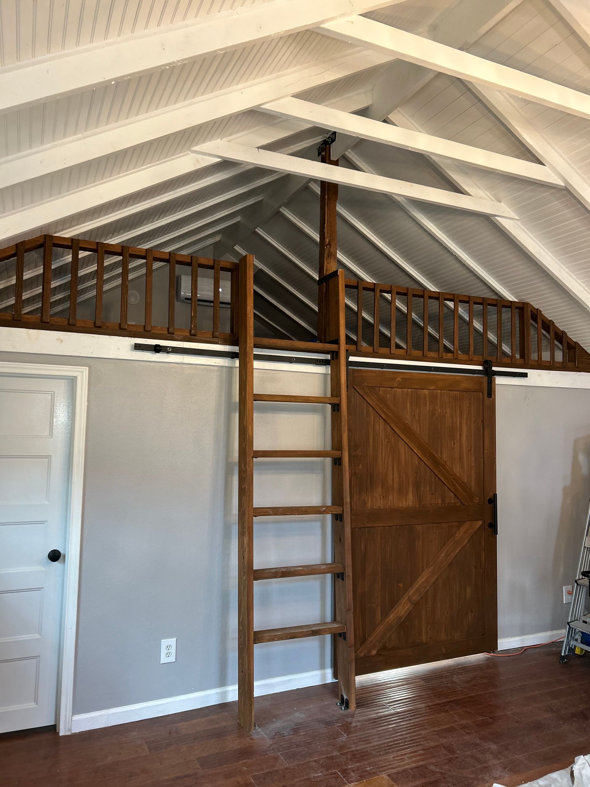 Interior loft with white vaulted ceiling, wooden ladder, and brown barn-style door over hardwood floor