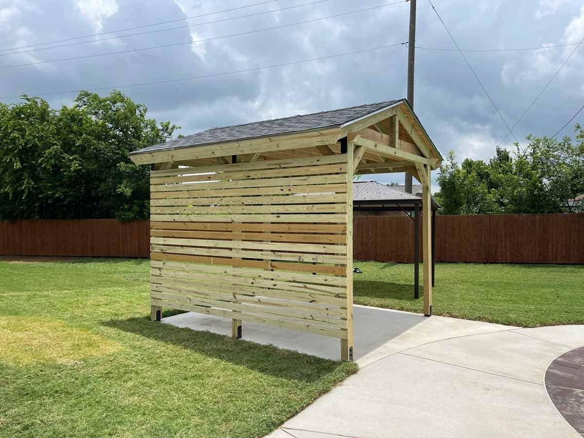 Small wooden shelter beside a sidewalk in a grassy yard, with a brown fence and cloudy sky.