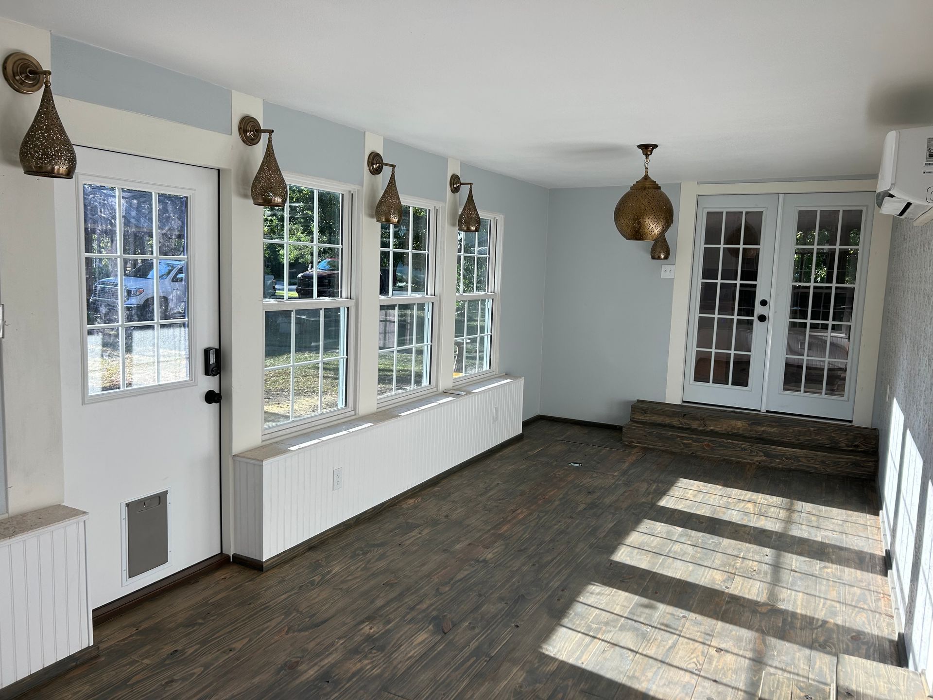Empty sunroom with white walls, large windows, and dark wood floors lit by sunlight.