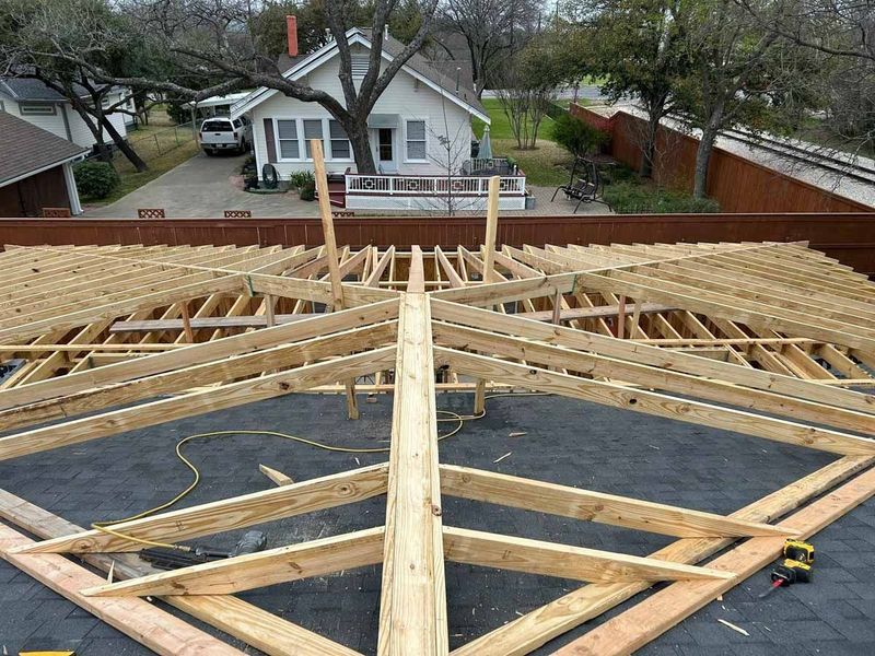 Roof framing under construction on a house, with exposed wooden trusses and a white home in the background.