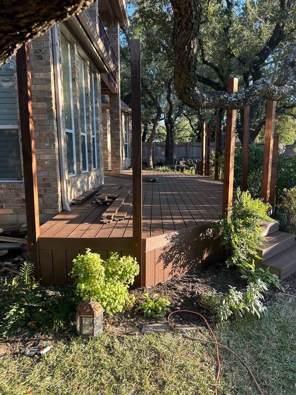 Wooden deck beside a glass-walled house, surrounded by trees and shrubs in a sunny yard