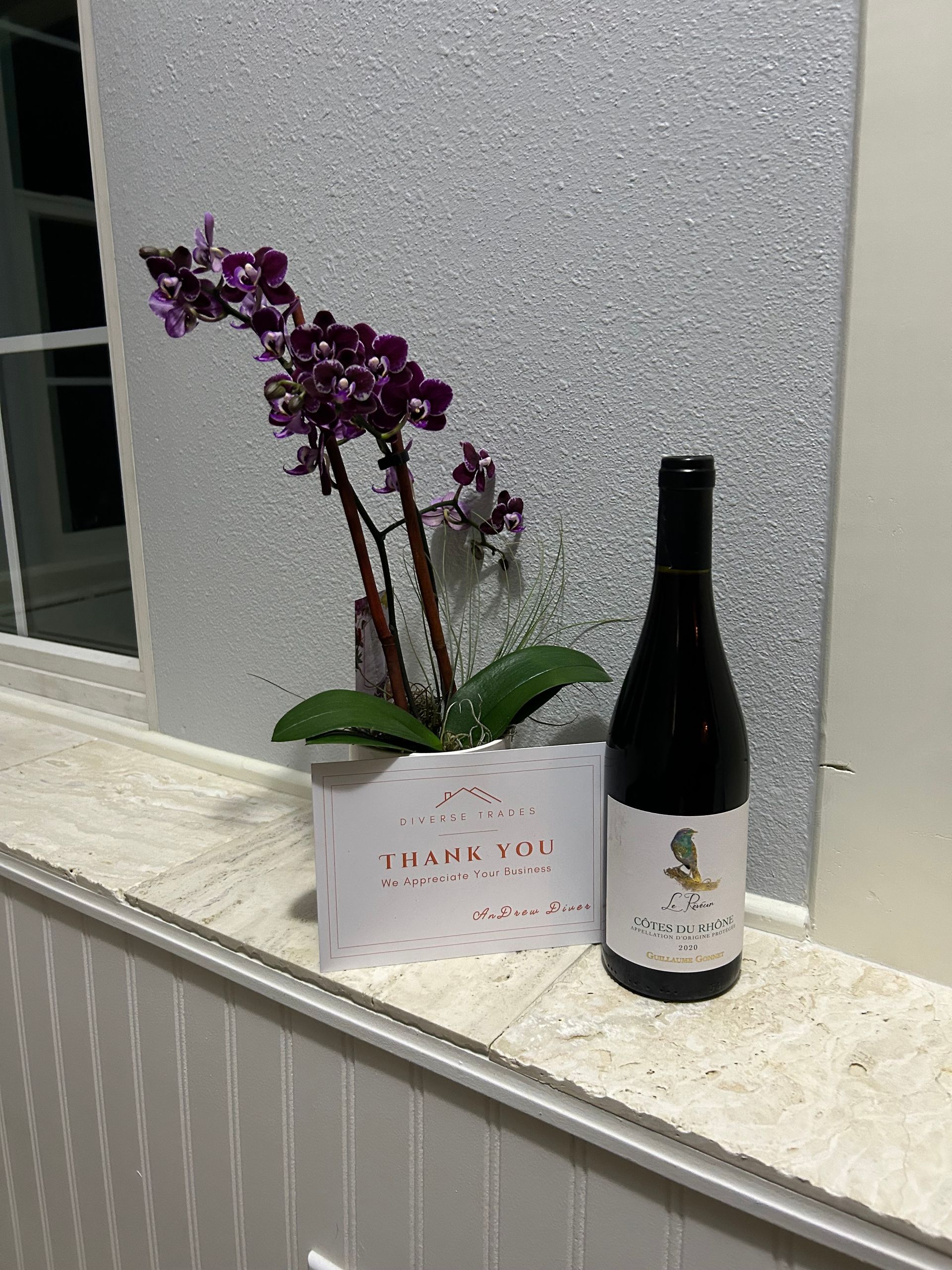 Purple orchid in a white box beside a dark wine bottle on a windowsill.