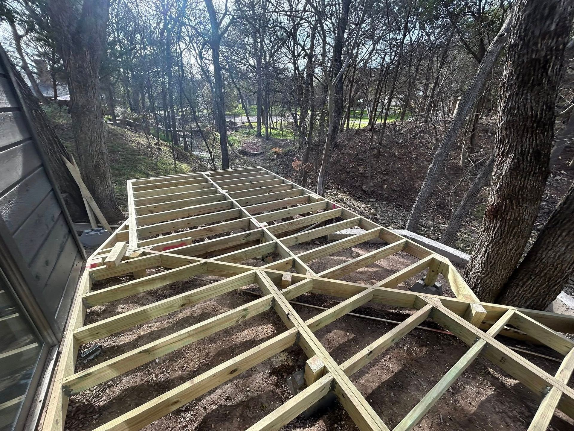 Wooden deck framing under construction beside trees, with beams laid over a sloped ground area.