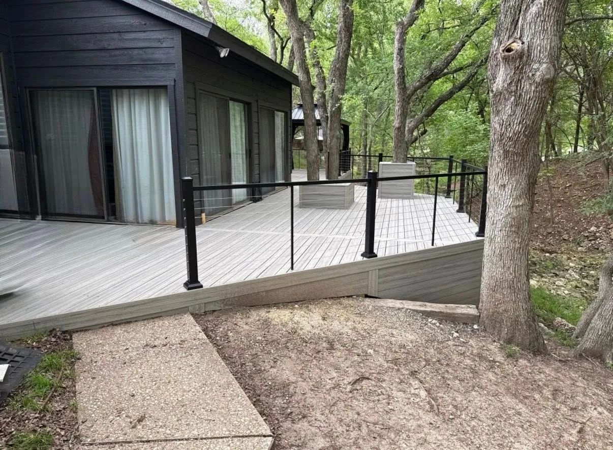 Black cabin beside a raised wooden deck with railings in a wooded area