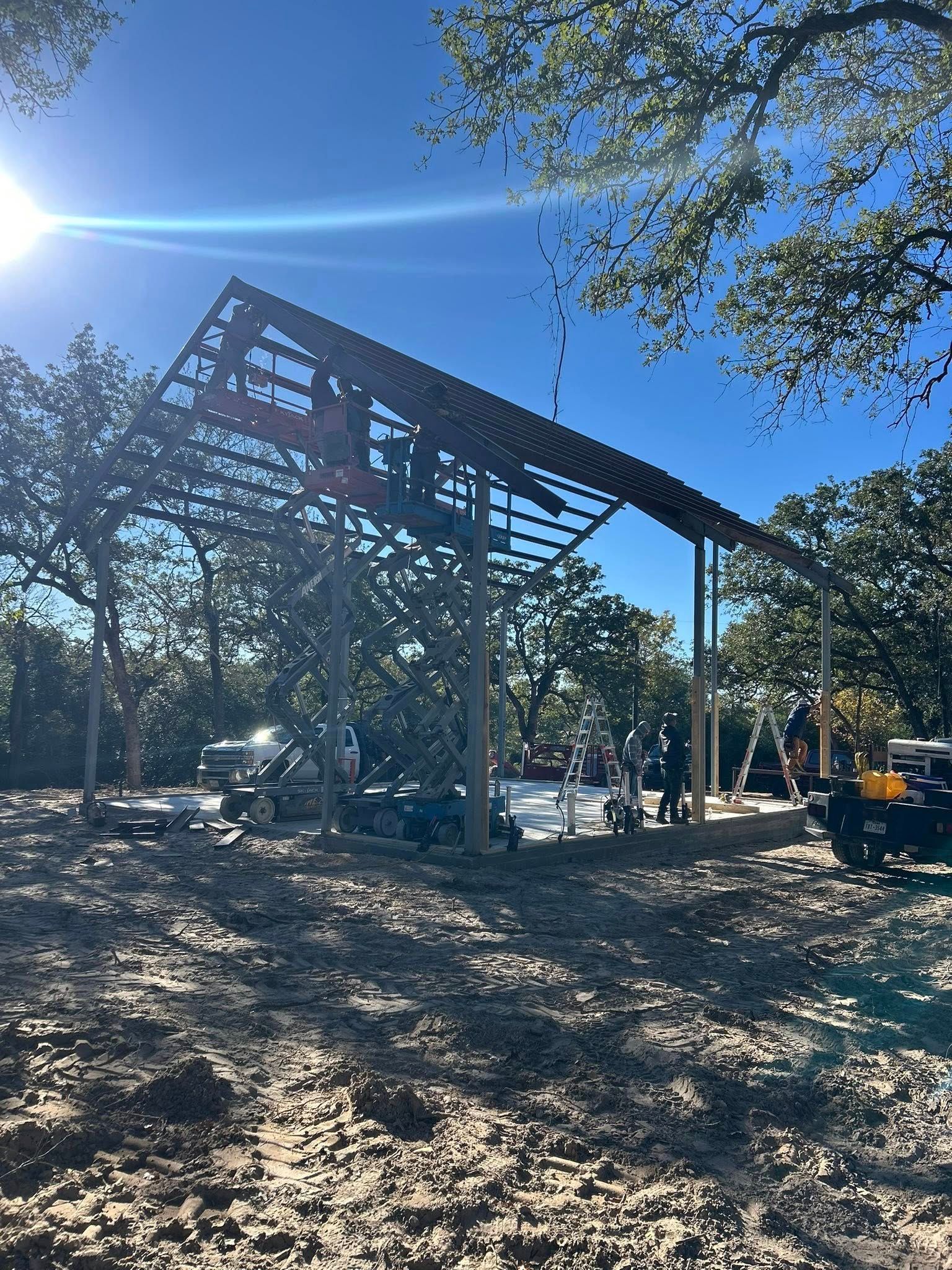Steel-framed building under construction on a sunny wooded site, with a loader nearby
