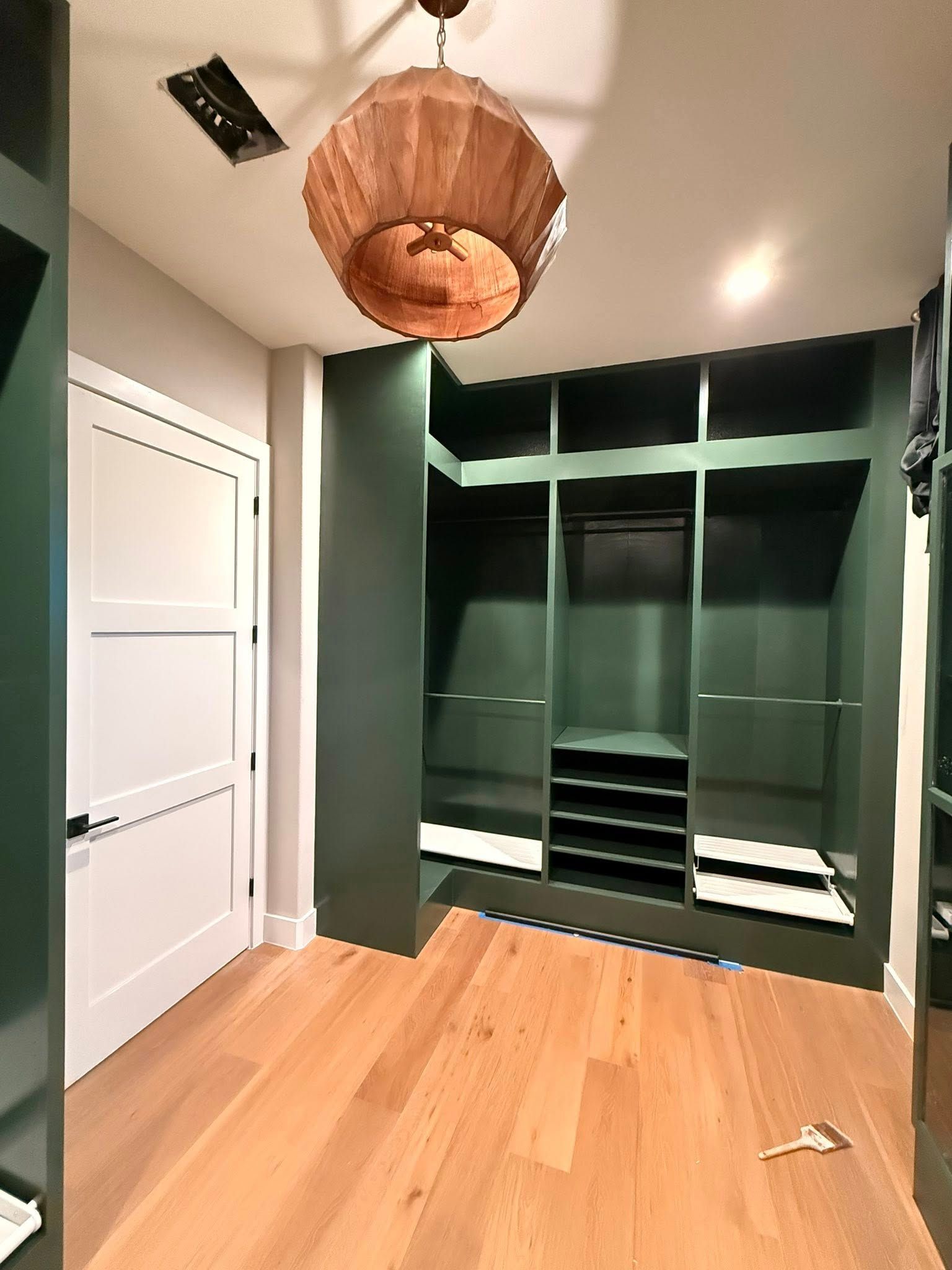 Modern walk-in closet with dark built-in shelves, white door, wood floor, and wicker pendant light