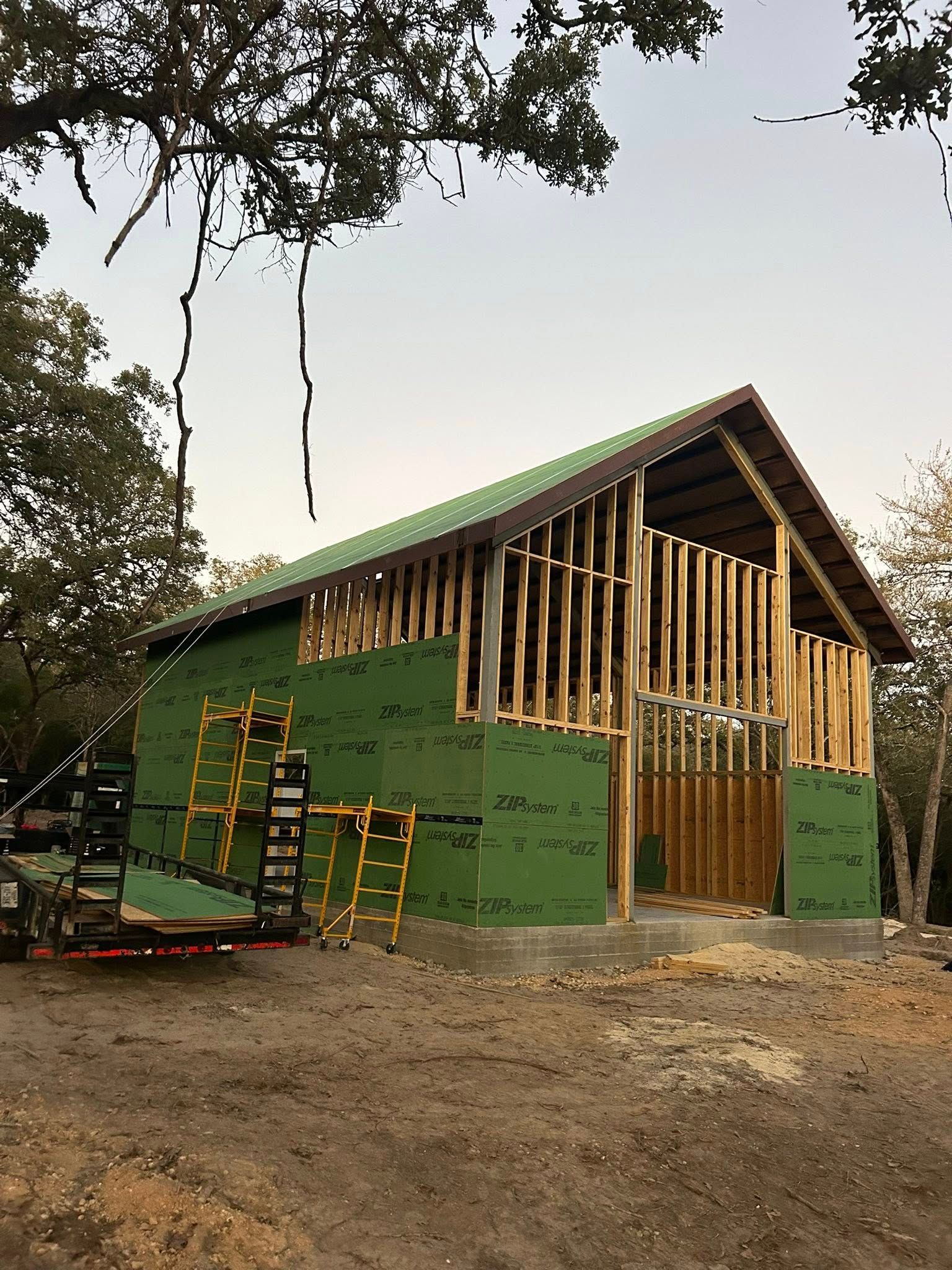 Green house under construction with exposed wooden framing and a ladder on the left, outdoors on bare ground.