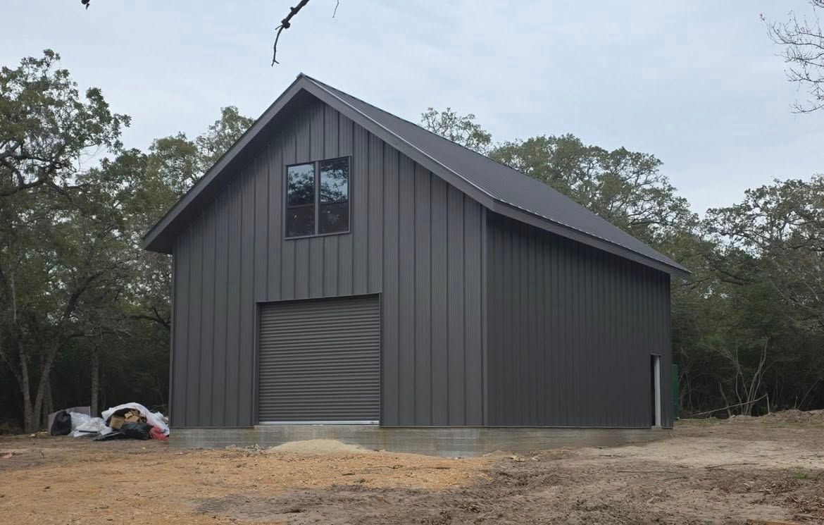 Gray metal barn with a large roll-up door and small upper window in a wooded clearing.