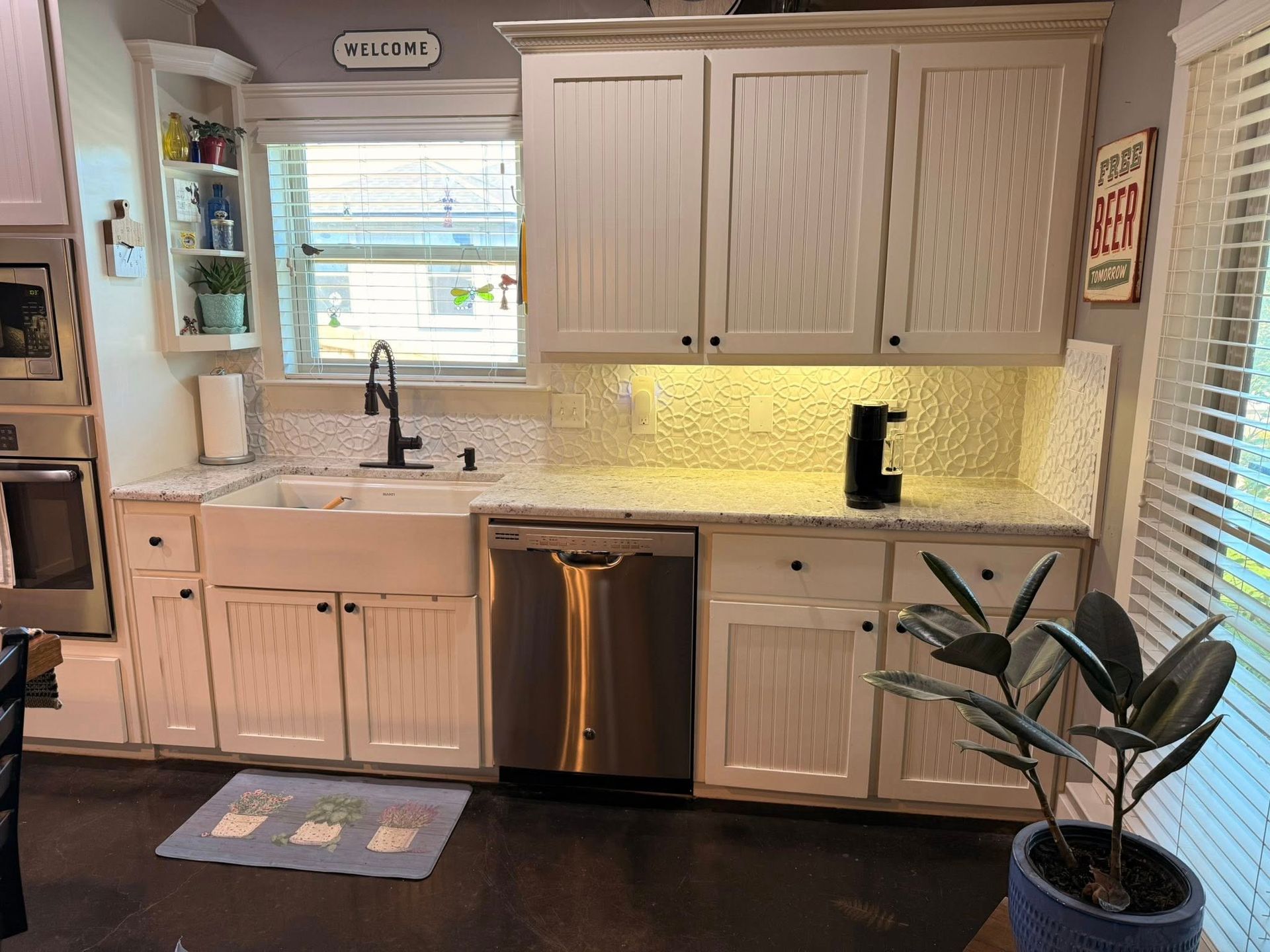 Kitchen with light wood cabinets, stainless dishwasher, sink under window, and a potted plant by the counter.