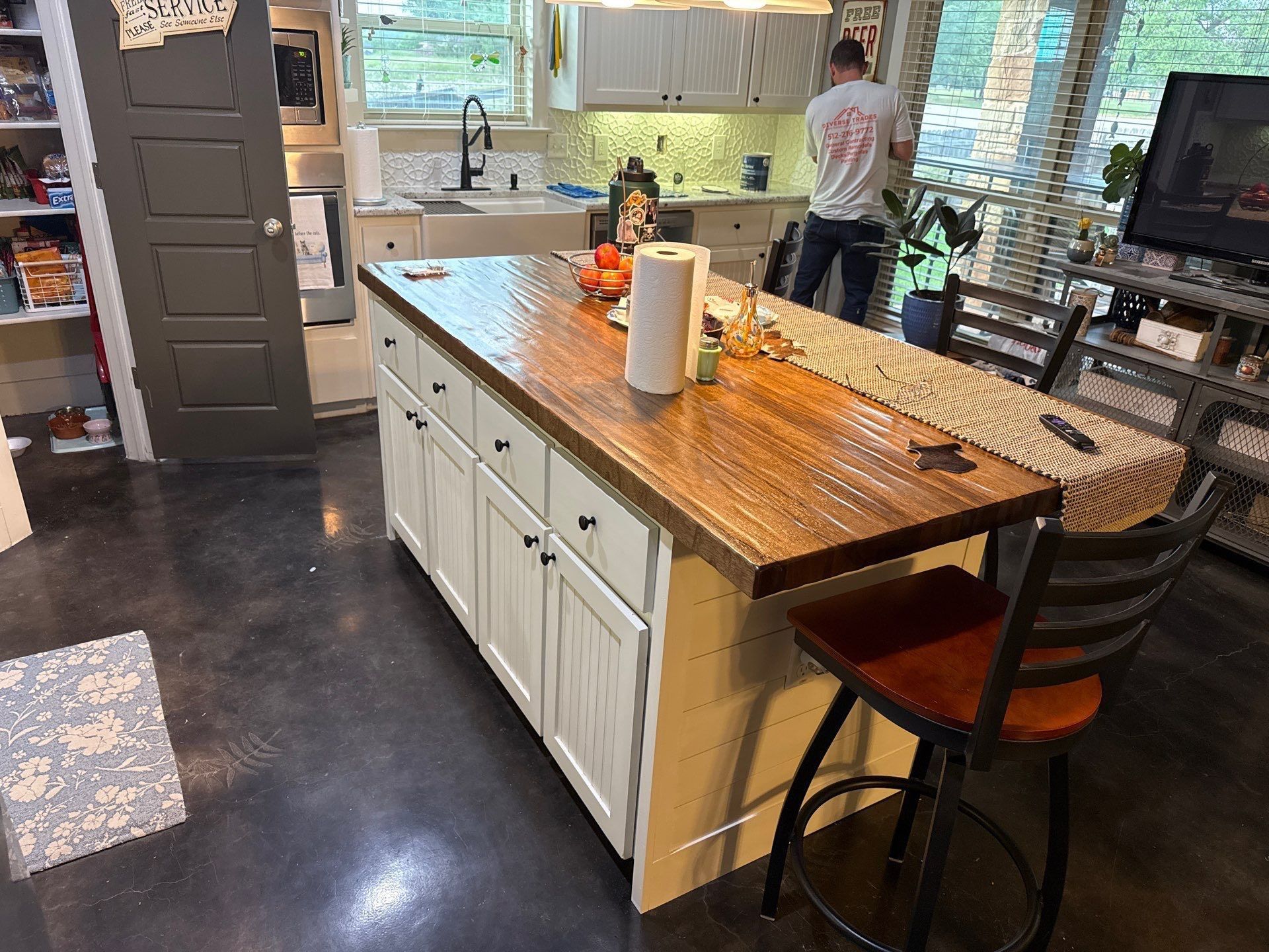 Kitchen with a wooden island, white cabinets, black floor, and a person standing by the sink near a window.