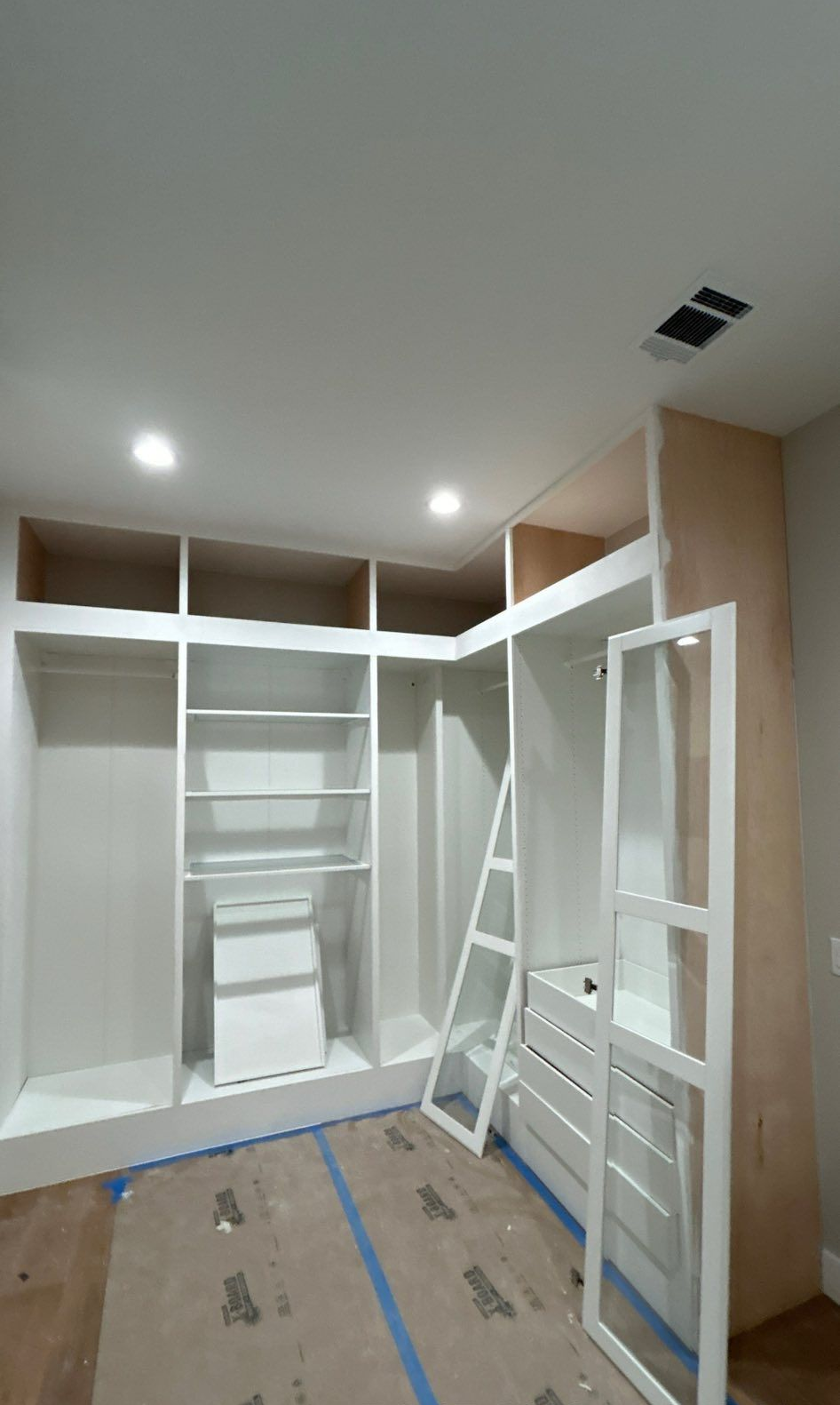 Built-in white closet under construction with shelves, doors, and tools in a room with unfinished flooring.