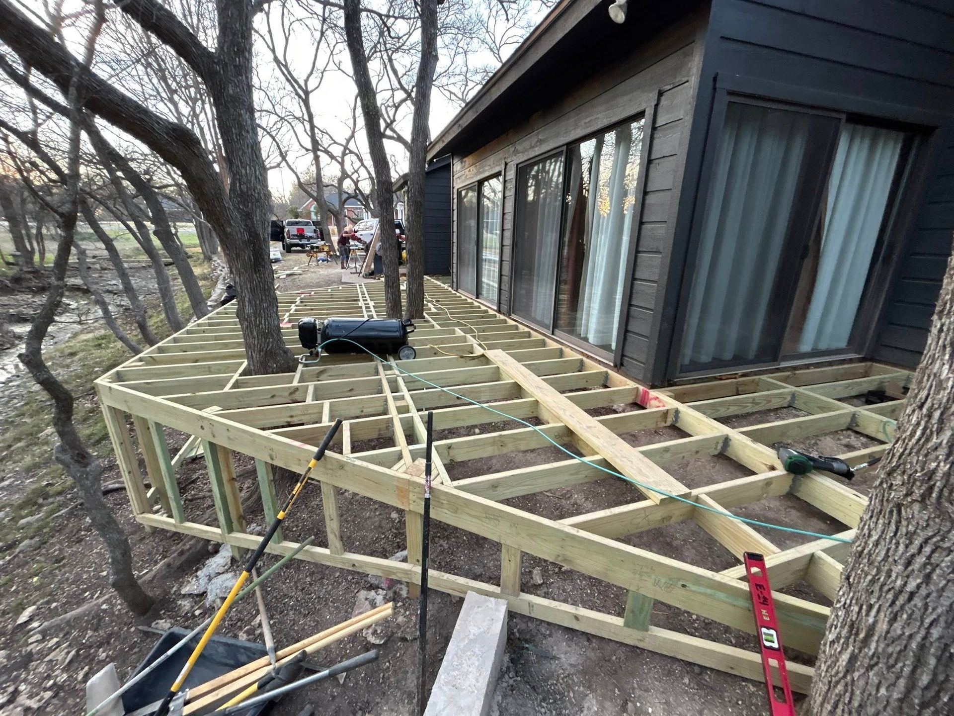 Wooden deck framing under construction beside a house, with tools and support posts on a wooded slope.