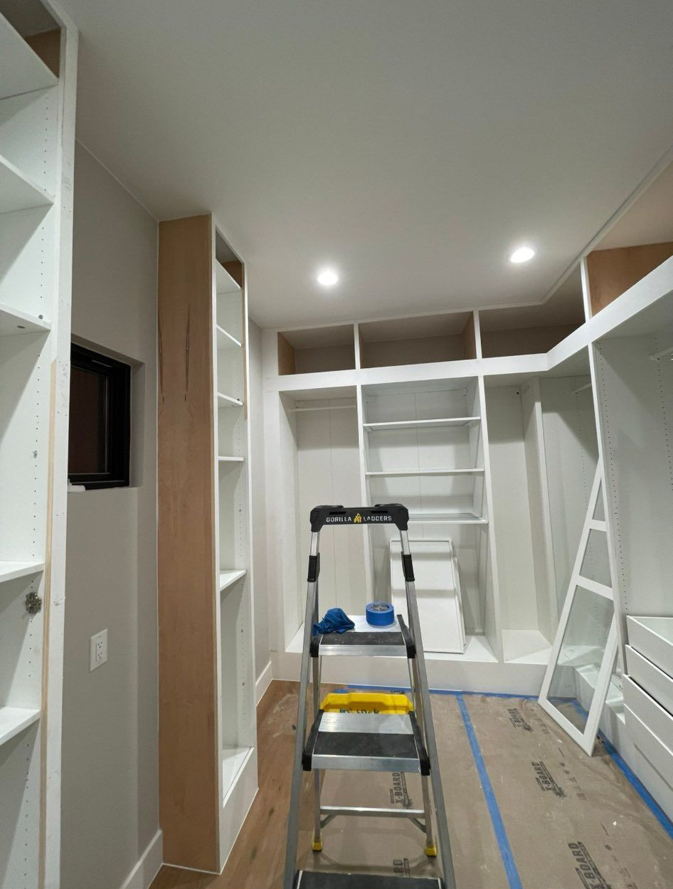 Narrow walk-in closet under renovation with ladder, white shelving, and blue painter’s tape on the floor