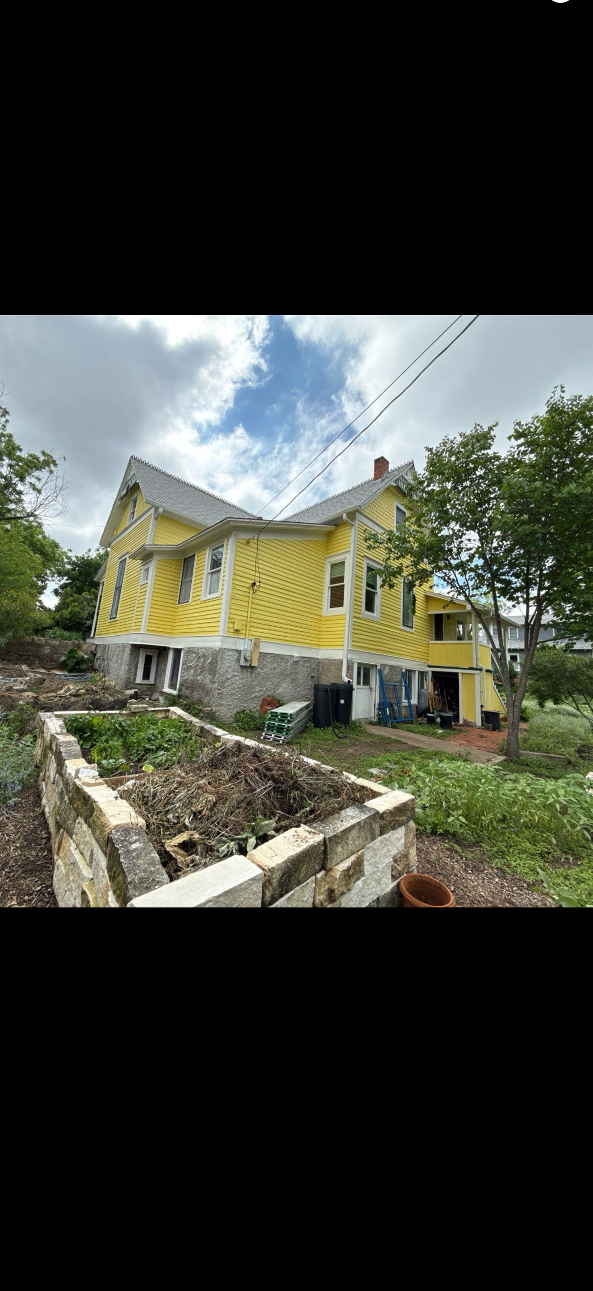 Yellow, weathered house with damaged roof beside a stone wall and trees under a cloudy sky