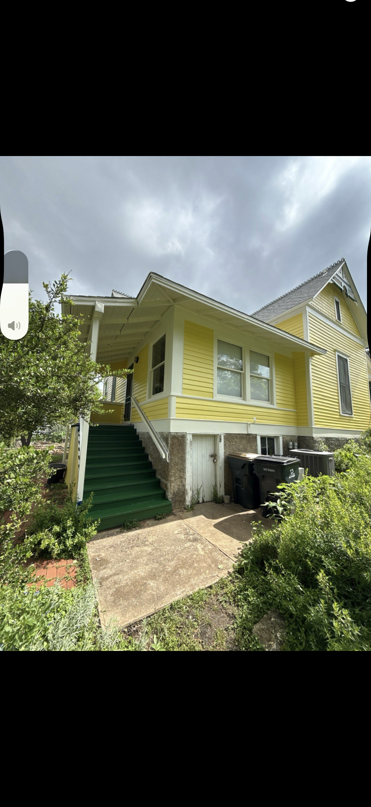Yellow house with white trim, green stairs, and a garden under a cloudy sky