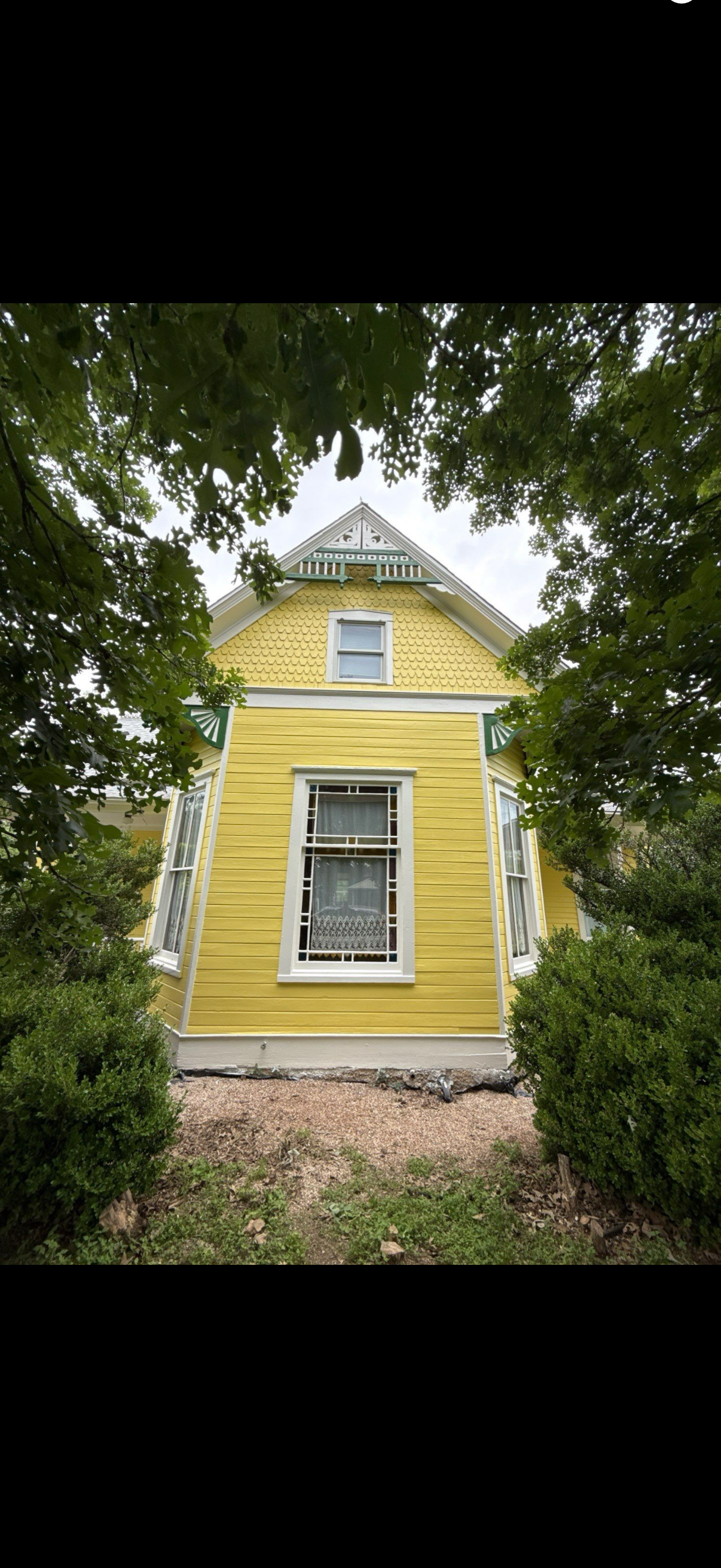 Yellow Victorian house viewed through trees, with ornate white trim and a front window.