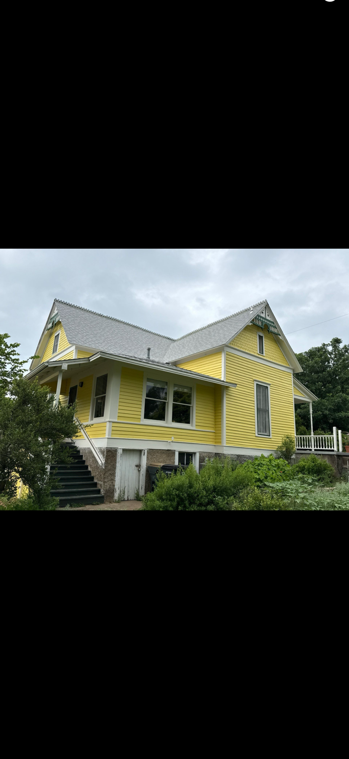 Yellow wooden house with a steep gray roof and front porch, surrounded by greenery