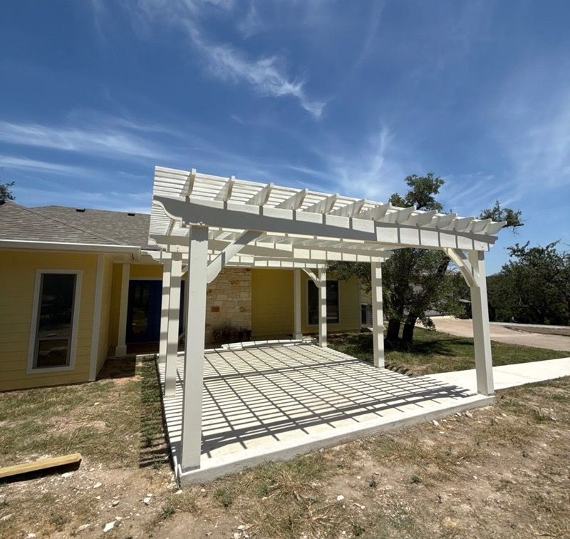 White pergola attached to a yellow house, casting shadows on a concrete patio under a blue sky.
