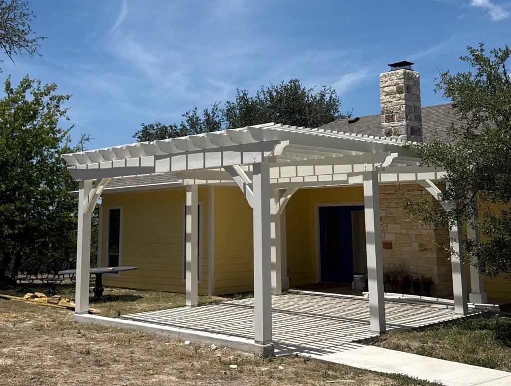 Yellow house with a white pergola-covered patio and stone chimney, surrounded by dry grass and trees