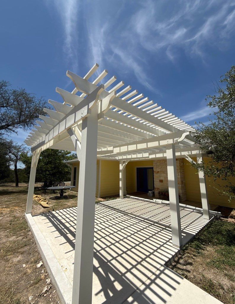 White pergola-covered patio beside a yellow house under a bright blue sky with shadows on the floor