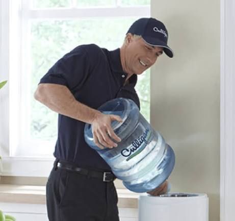 Man pouring water from a large bottle into a dispenser near a window