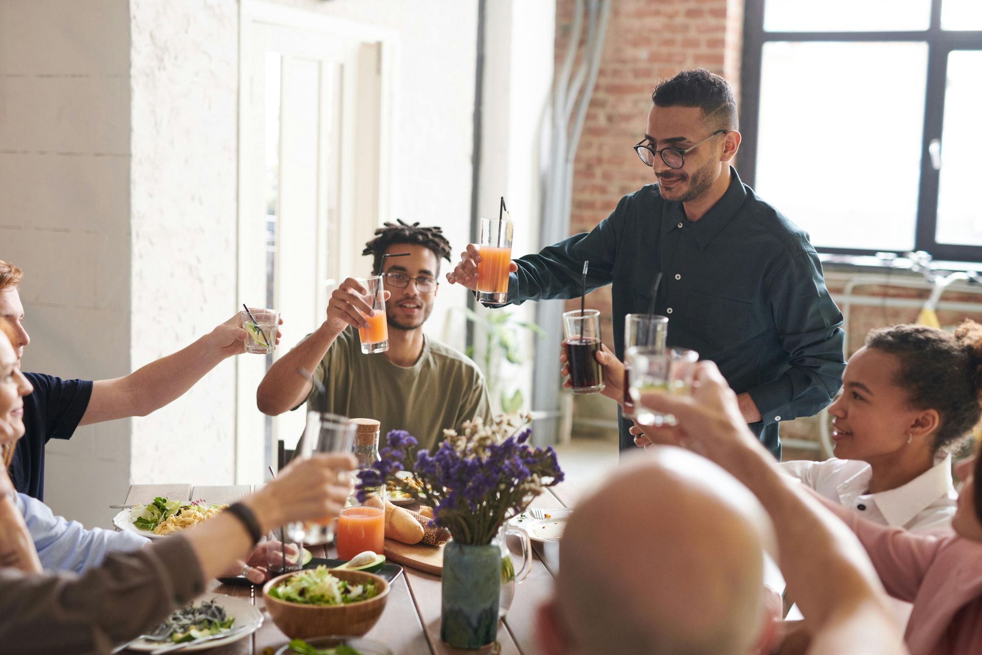Friends at a table, toasting with drinks, celebrating; bright, sunny room, diverse group.