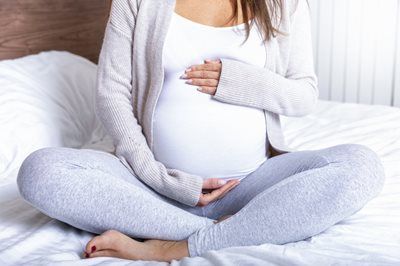 Pregnant person sitting cross-legged on a bed, resting hands on a rounded belly.