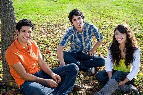 Three friends sitting on grass outdoors, smiling in the sun.