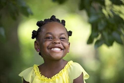 Smiling child in a yellow shirt outdoors with green foliage in the background