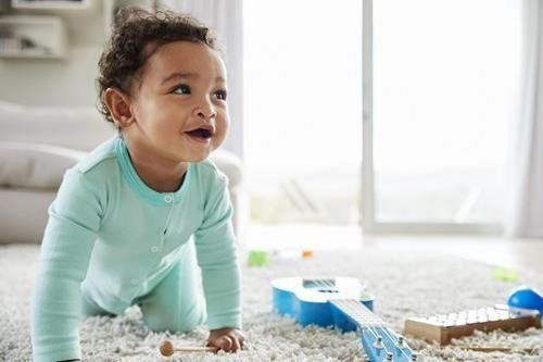 Baby in a mint-green onesie crawling on a fluffy white rug near toys by a bright window