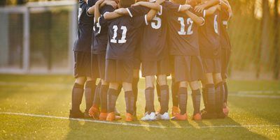 Soccer team huddled together on a sunlit field, wearing dark jerseys and shorts.