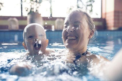 Adult and baby smiling in a swimming pool, splashing water indoors