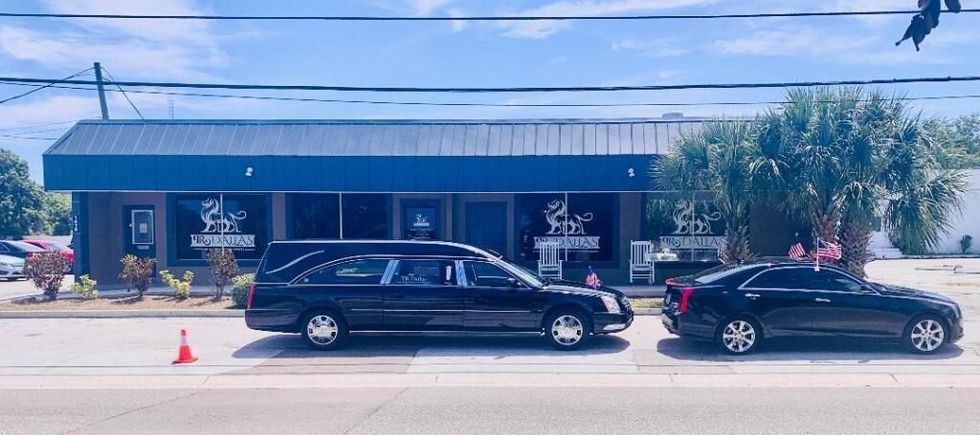 Black stretch limousine and black sedan parked outside a blue building with palm trees and cones nearby