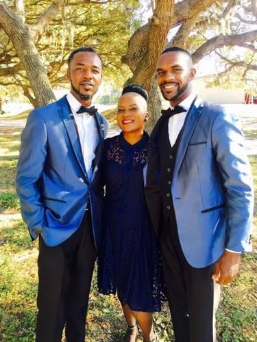 Three people smiling outdoors under a tree, wearing formal blue outfits.