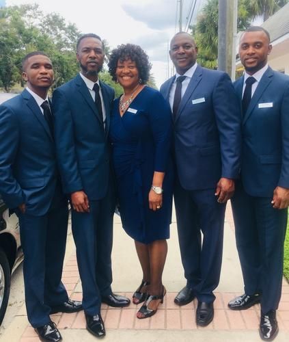 Five people in matching navy suits posing outdoors on a walkway.
