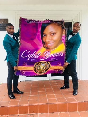 Two people holding a large purple memorial banner with a portrait, standing on a tiled porch.