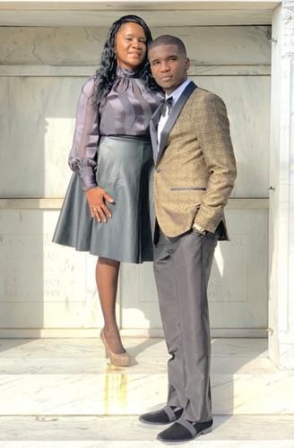 Couple posing together outdoors against a light wall, wearing coordinated formal outfits.