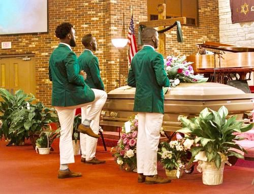 Funeral pallbearers in green jackets carry a casket in a church, surrounded by flowers.